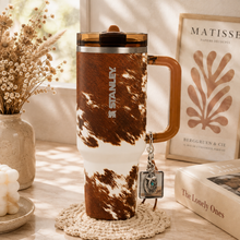 A Stanley insulated tumbler with a brown and white cowhide pattern, a translucent brown handle, and a metal keychain, displayed on a table next to a vase of dried flowers and books.