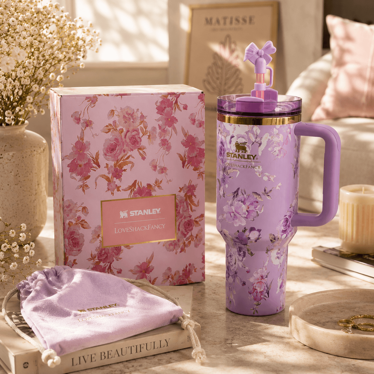A lavender floral Stanley tumbler with a purple handle and matching lid sits beside its pink floral product box on a styled tabletop. A lavender drawstring pouch, books, flowers, and soft home decor are visible in a warm, sunlit interior setting.