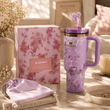 A lavender floral Stanley tumbler with a purple handle and matching lid sits beside its pink floral product box on a styled tabletop. A lavender drawstring pouch, books, flowers, and soft home decor are visible in a warm, sunlit interior setting.
