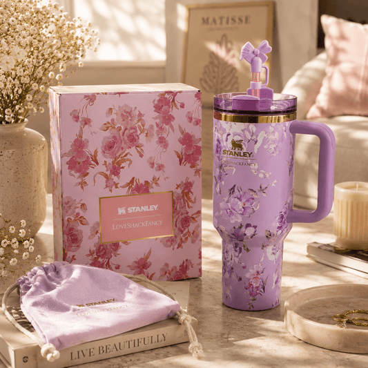 A lavender floral Stanley tumbler with a purple handle and matching lid sits beside its pink floral product box on a styled tabletop. A lavender drawstring pouch, books, flowers, and soft home decor are visible in a warm, sunlit interior setting.