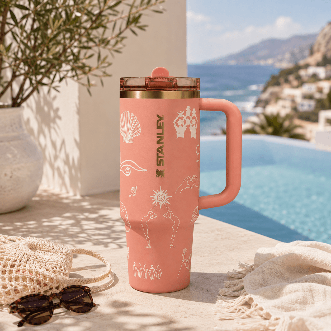 A pink Stanley travel mug with white line-art illustrations and a clear lid, set on a stone patio next to a swimming pool with a Mediterranean background.