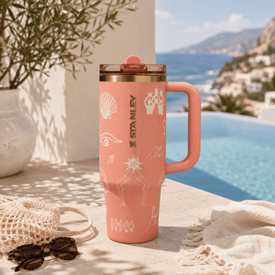 A pink Stanley travel mug with white line-art illustrations and a clear lid, set on a stone patio next to a swimming pool with a Mediterranean background.