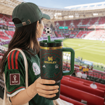 Person holding a Stanley mug with a soccer-themed design in a stadium setting