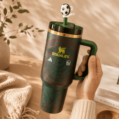Green Stanley mug with a soccer-themed lid held by a hand on a wooden surface.